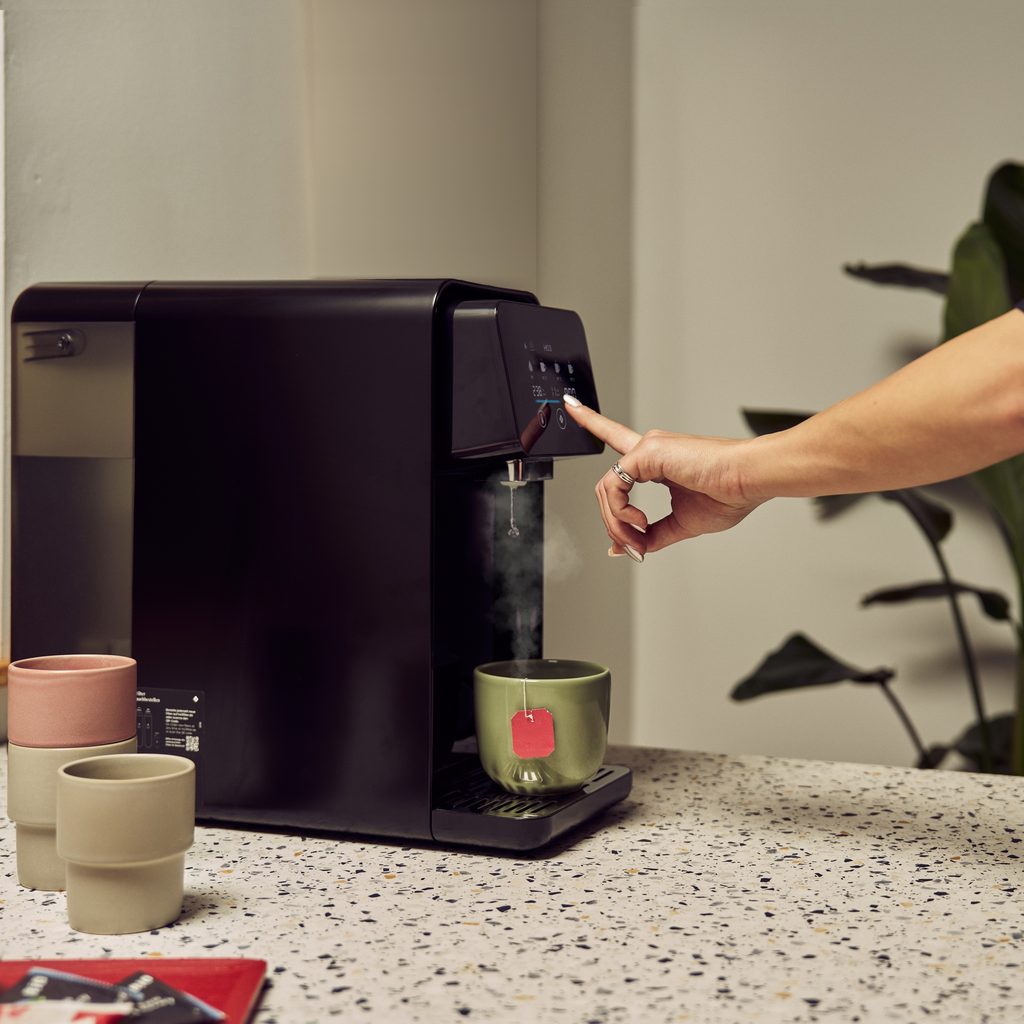Hand drückt am Home One auf Heißwasser, dampfendes Wasser läuft in eine Teetasse